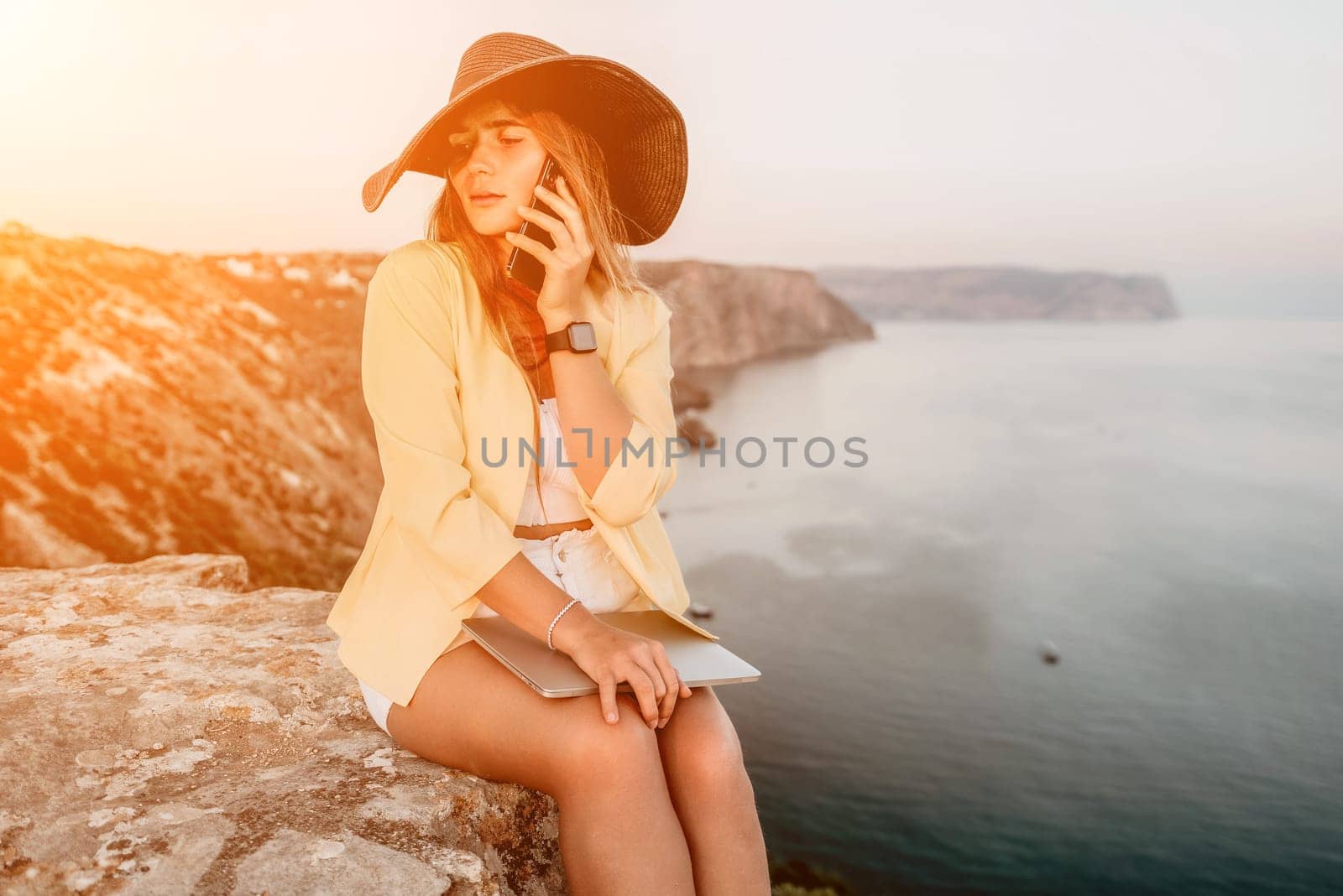 Successful business woman in yellow hat working on laptop by the sea. Pretty lady typing on computer at summer day outdoors. Freelance, travel and holidays concept.