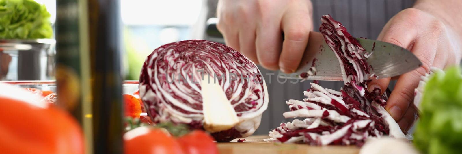 Closeup of a male cook hand using knife slicing fresh red cabbage on cutting board in kitchen at home. Cooking vegetable salad concept