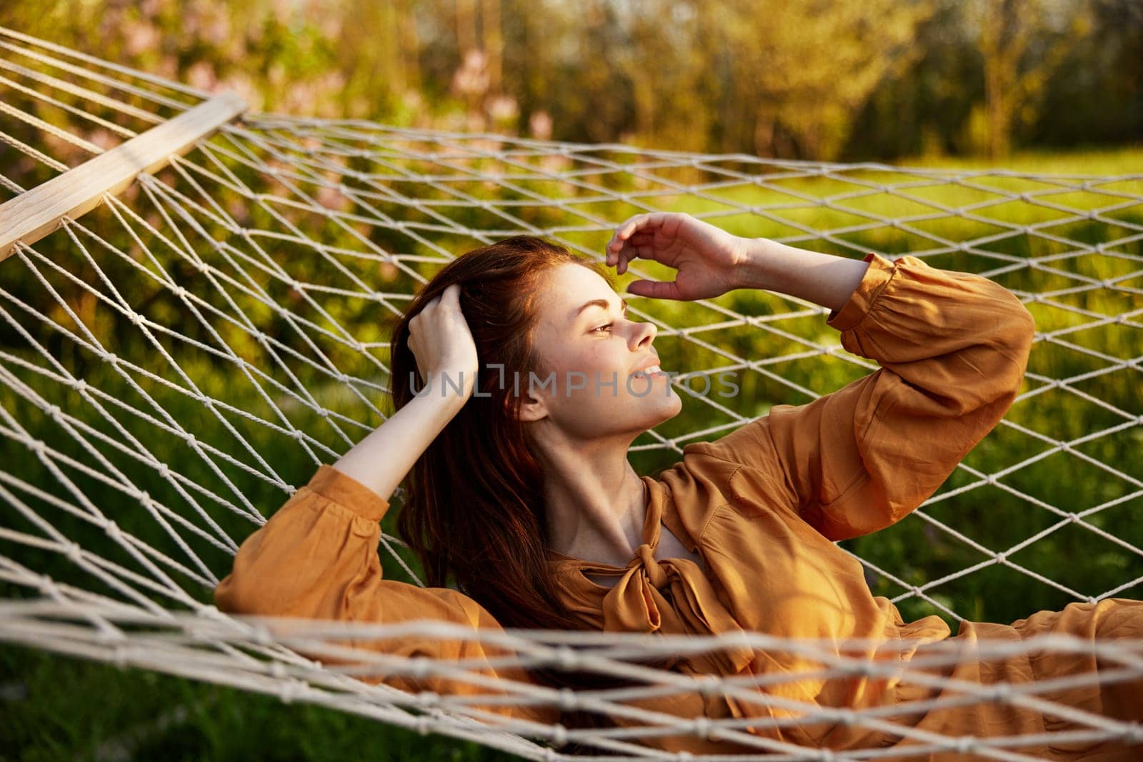 a happy woman is resting in a mesh hammock with her hands behind her head, straightening her hair, smiling happily looking away, enjoying a sunny day. High quality photo