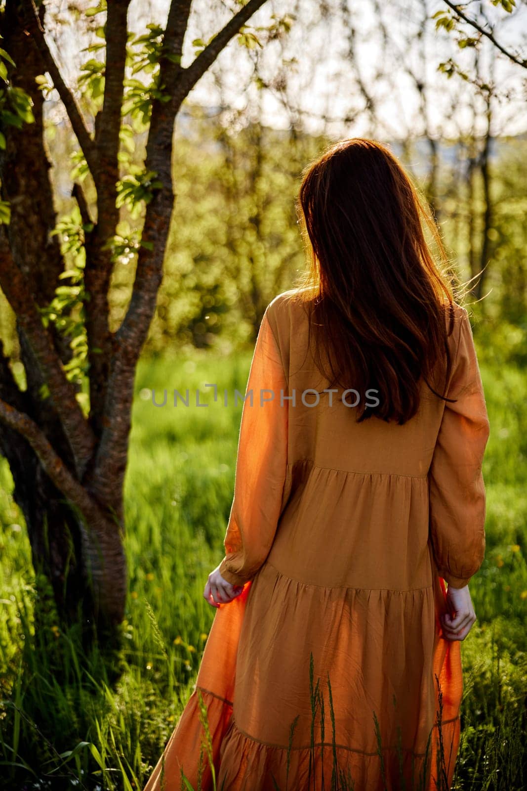 a lovely woman stands in nature in a bright, long orange dress, illuminated from the back by the sunset rays of the sun and holds the skirt of the dress with her hand, looking to the side enjoying the day. High quality photo