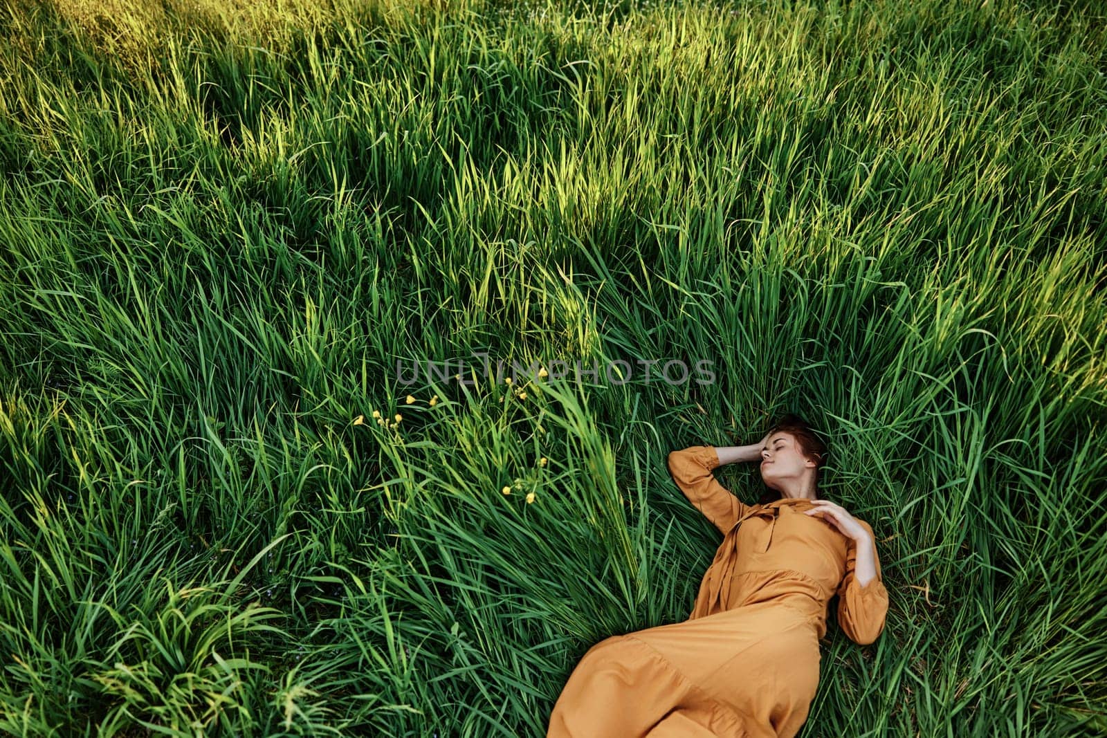 a sweet, calm woman in an orange dress lies in a green field enjoying the silence and peace. Horizontal photo taken from above by Vichizh