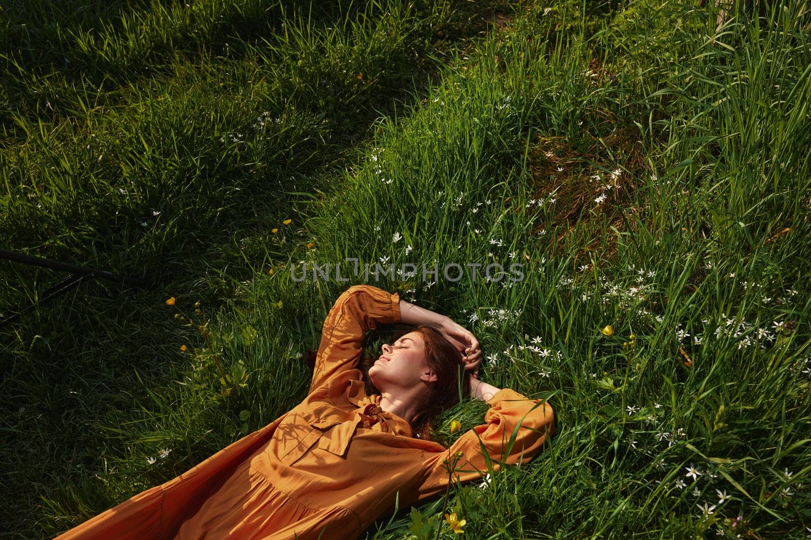 horizontal photo of a happy, relaxed woman, resting lying in the grass, in a long orange dress, with closed eyes and a pleasant smile on her face, enjoying harmony with nature and recuperating by Vichizh