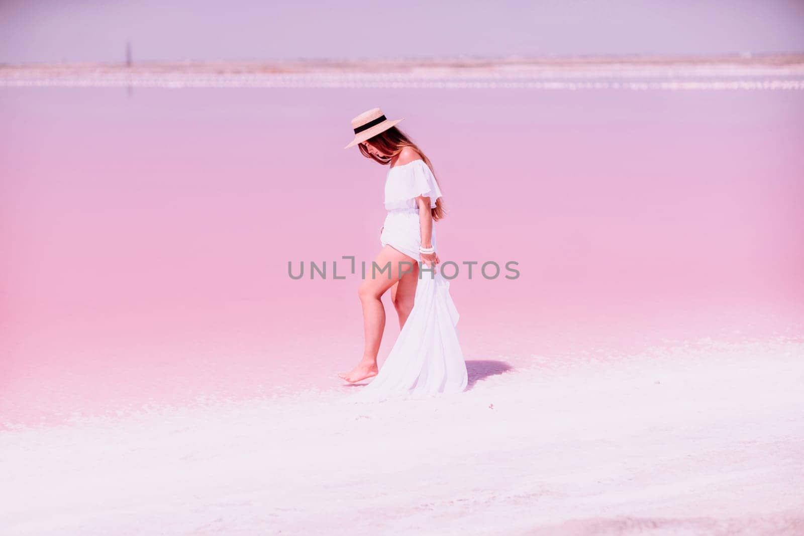 Woman in pink salt lake. She in a white dress and hat enjoys the scenic view of a pink salt lake as she walks along the white, salty shore, creating a lasting memory