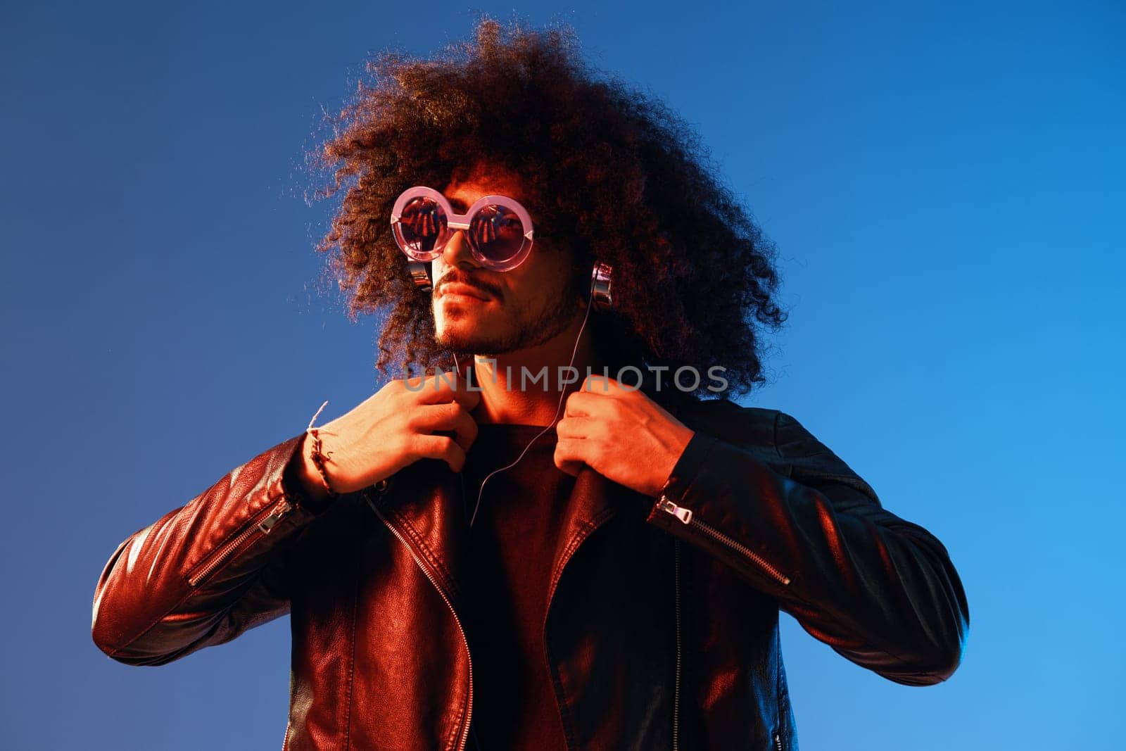 Portrait of a stylish man with curly hair with glasses and headphones on a blue background multinational, colored light, black leather jacket trend, modern concept. High quality photo