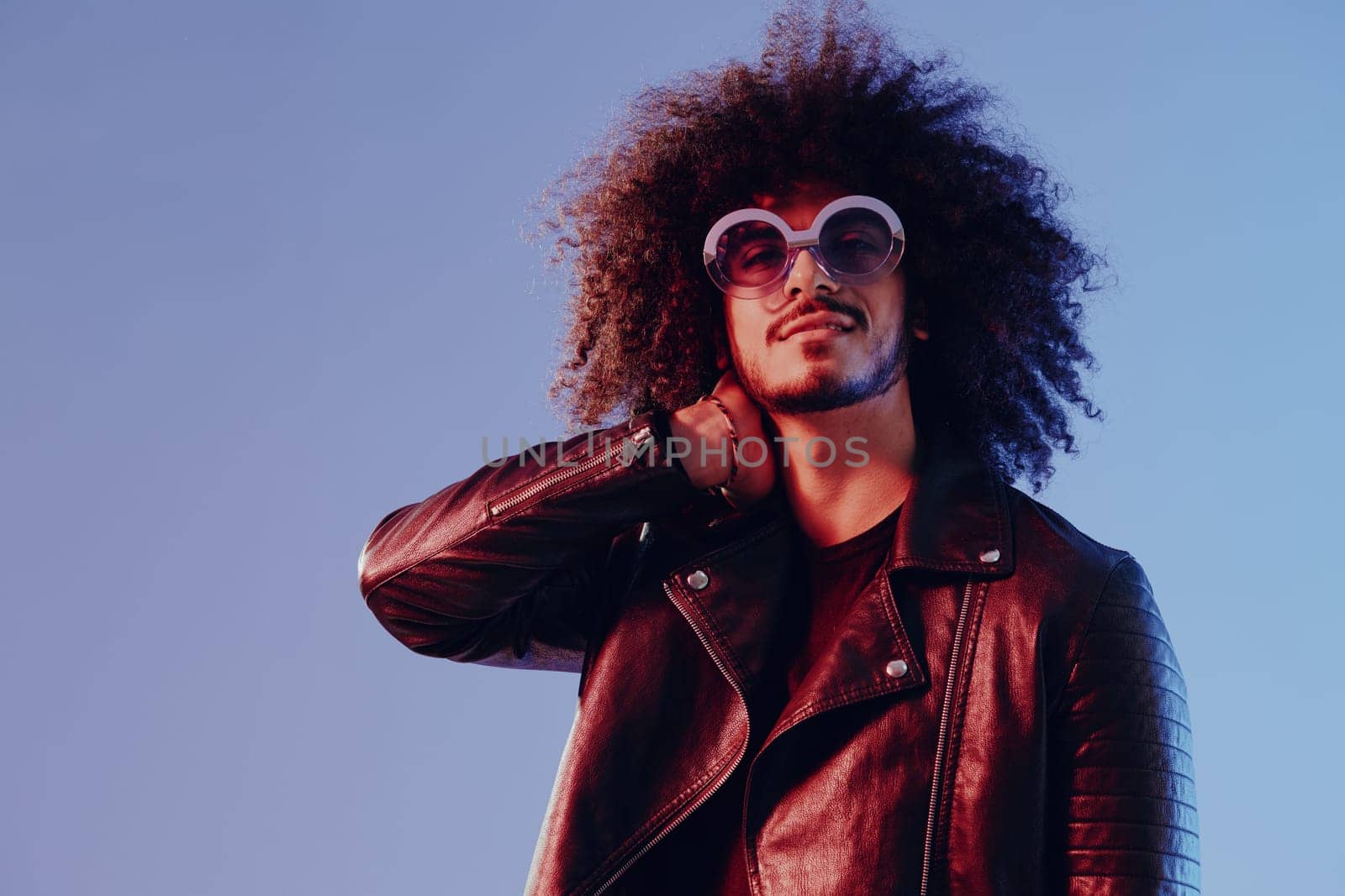 Portrait of a stylish man with curly hair with glasses smile on blue background multinational, color light, black leather jacket trend, modern concept. by SHOTPRIME