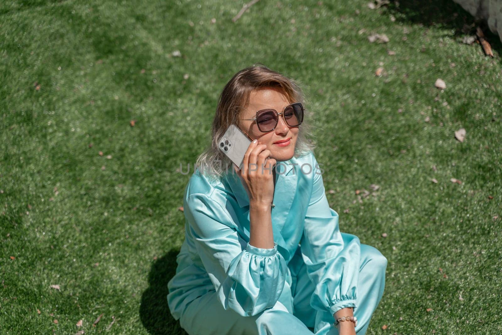 Woman grass. Happy woman with blond curly hair in glasses and in a blue overalls sits on the summer fresh green grass with a phone in her hands. by Matiunina