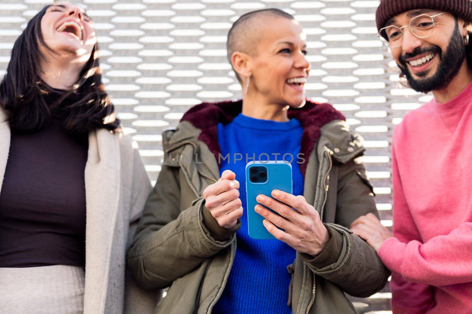 three young friends laughing and enjoying the moment using together a mobile phone, focus on the phone, concept of technology of communication and modern lifestyle
