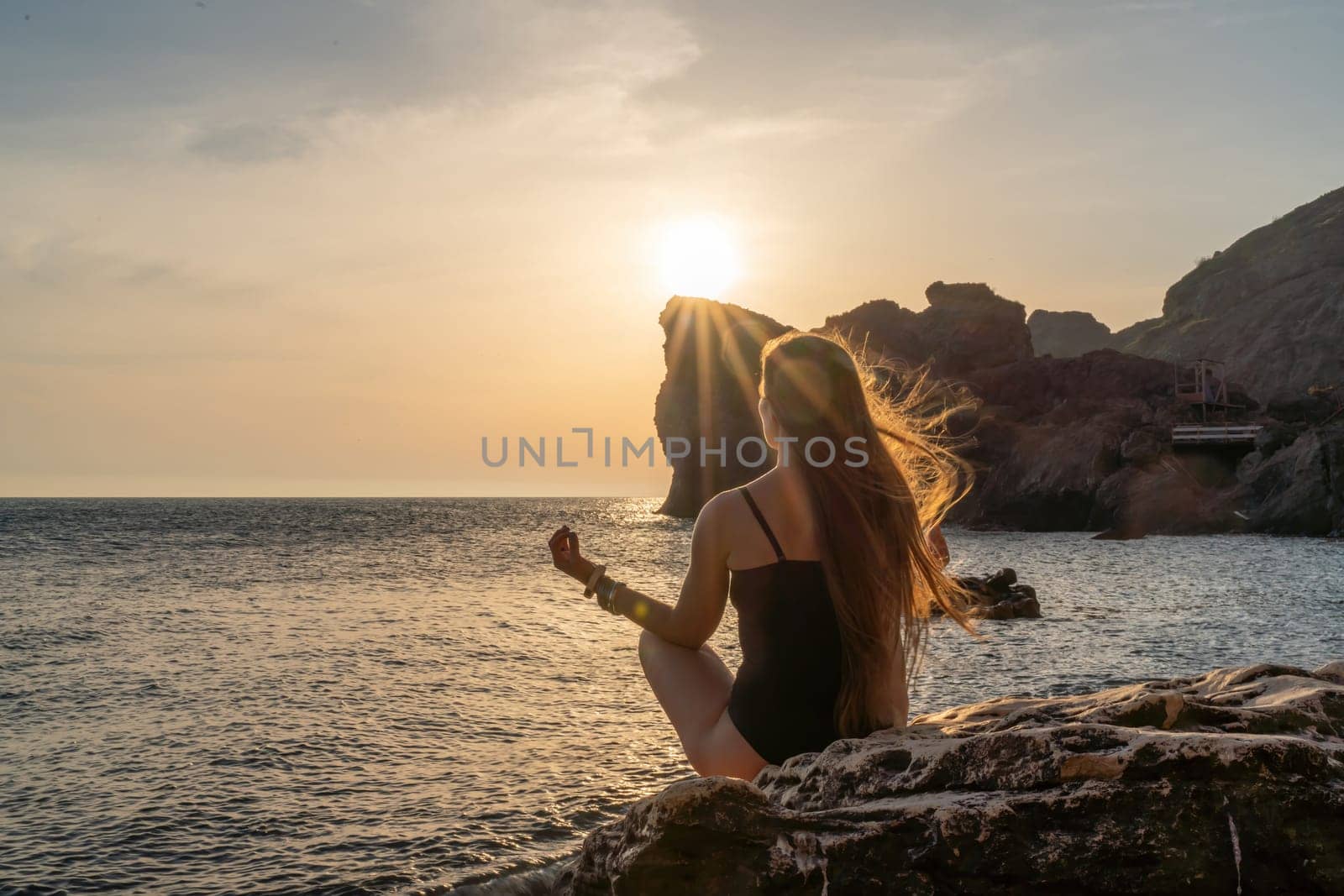 Woman tourist enjoying the sunset over the sea mountain landscape. Sits outdoors on a rock above the sea. She is wearing jeans and a blue hoodie. Healthy lifestyle, harmony and meditation by Matiunina