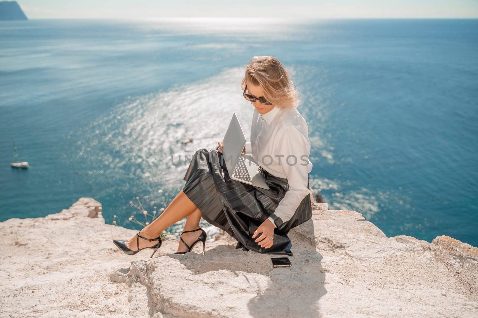 Business woman on nature in white shirt and black skirt. She works with an iPad in the open air with a beautiful view of the sea. The concept of remote work