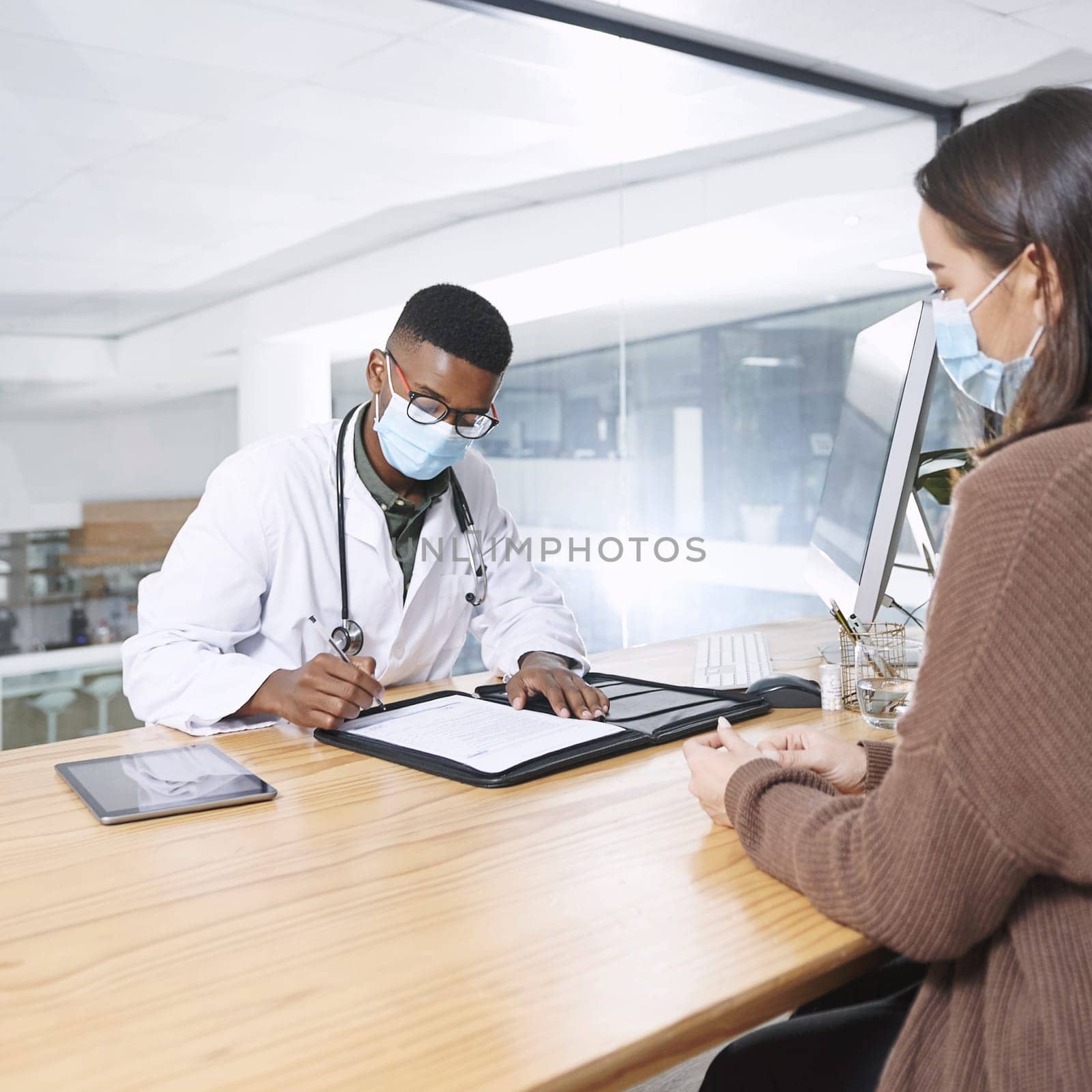 This is what I need you to get from the pharmacy. a young doctor sitting with his patient and wearing a mask while writing a prescription during a consultation