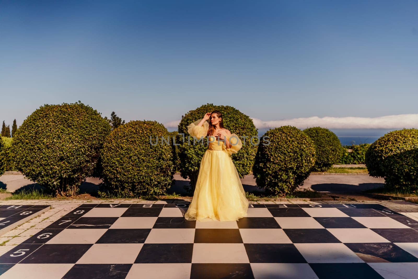 Woman yellow dress chess. A beautiful woman in a long puffy yellow dress poses on a chessboard in the park