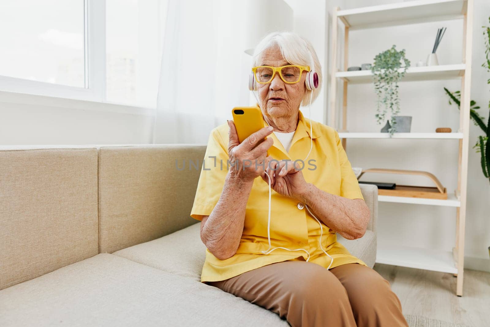 Happy elderly woman looking into her phone video call with headphones smile, technology for communication, bright modern interior, lifestyle online communication. High quality photo