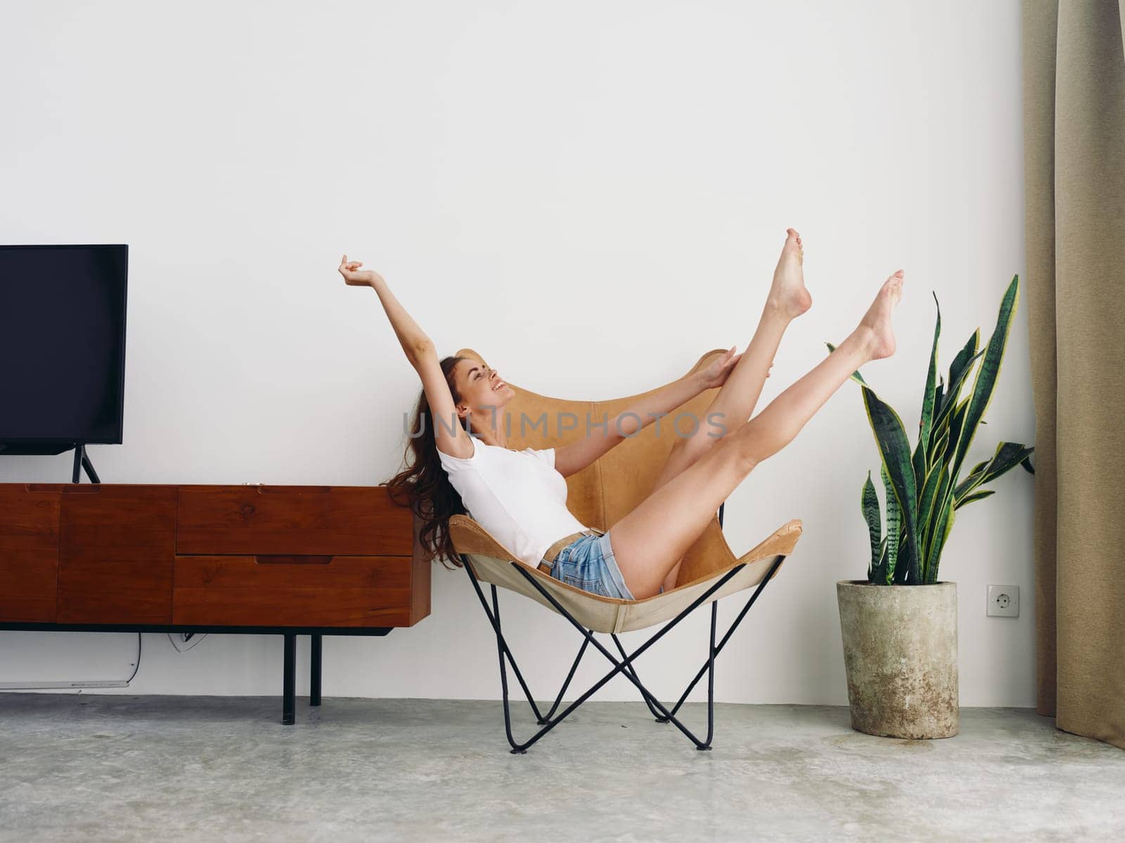 Woman lying in a leather armchair raised arms and legs cheerful happiness and a smile, relaxing at home stylish modern interior with white walls, copy space. by SHOTPRIME