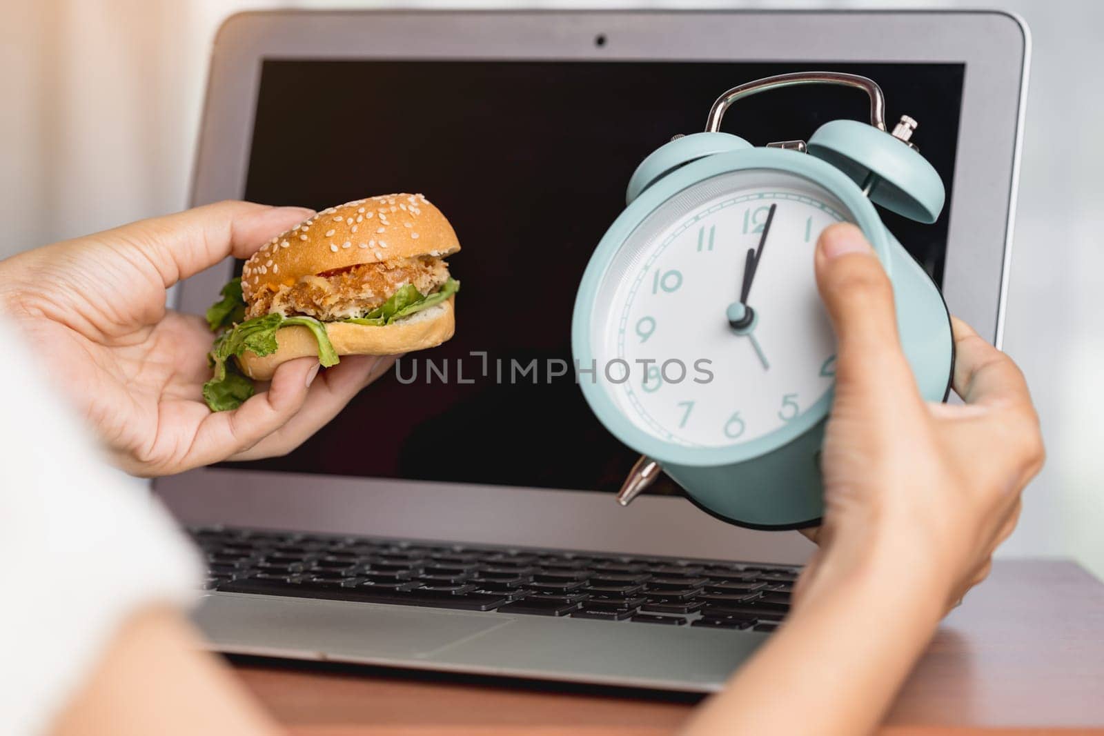Hand holding a delicious hamburger and alarm clock while working for the concept of eating and time management