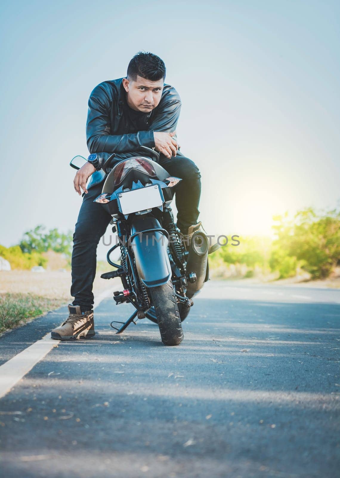 Handsome motorcyclist in jacket sitting on his motorcycle at the side of the road. Portrait of handsome biker on his motorbike looking at camera outdoors by isaiphoto