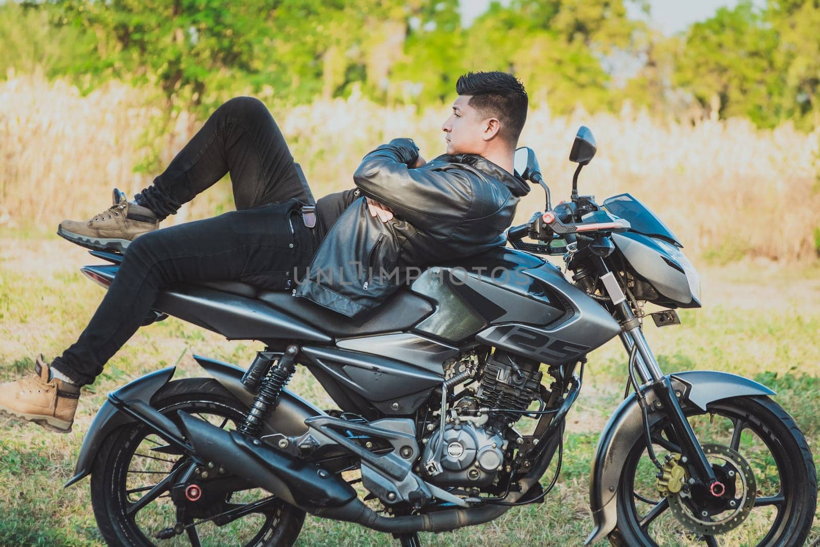 Biker man lying down on his motorbike outdoors. Motorcyclist in jacket lying on his motorcycle in the countryside
