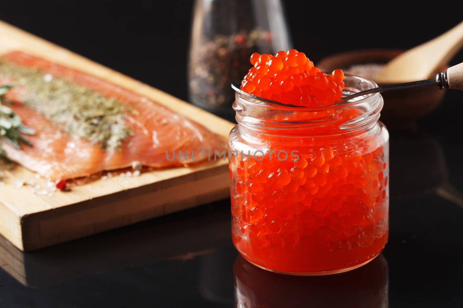 Salted fish fillet with spices, dill and rosemary on a wooden board and a jar of red caviar on a black background.
