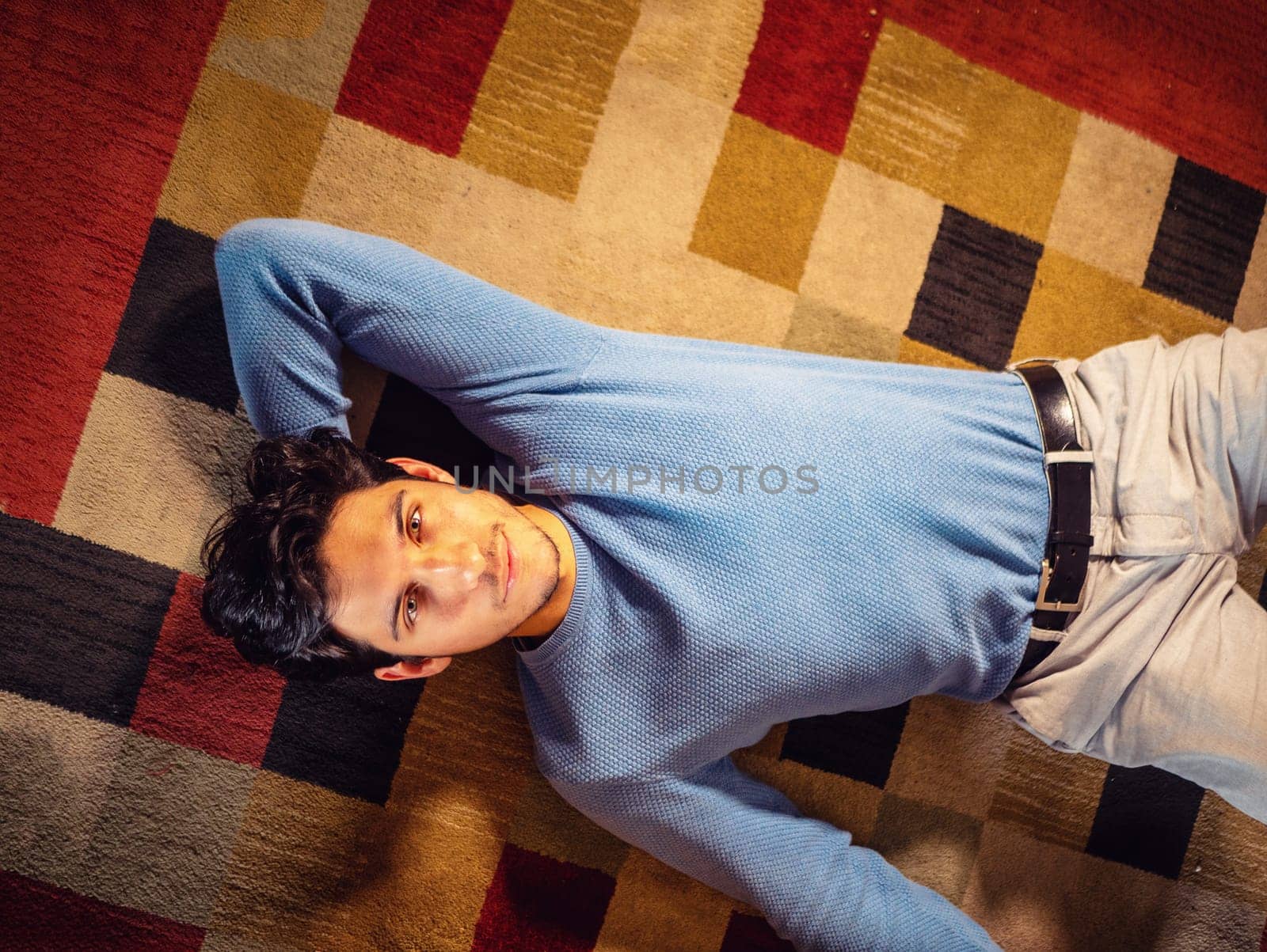 Cool dark-haired handsome young man laying on the floor over colorful rug, looking at camera, hand under his head