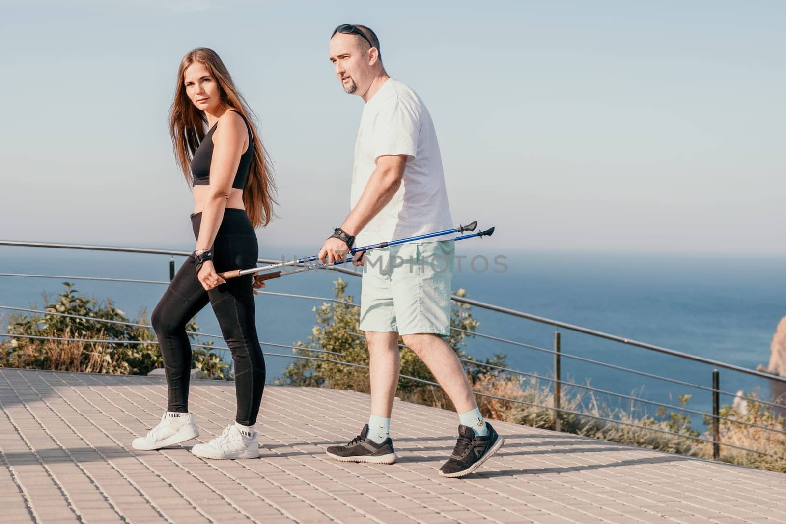 Happy Middle aged couple or friends practicing nordic walking in park near sea. Mature couple with trekking poles walking, practicing Nordic walking outdoors. Aging youthfully and sport concept.