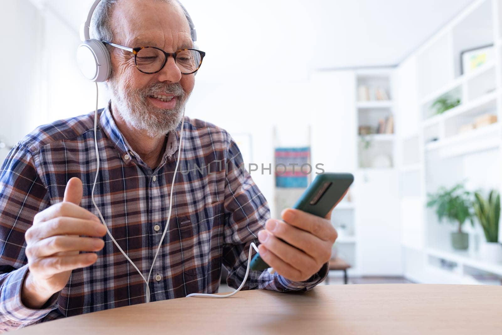 Senior man listening to music and dancing using mobile phone and headphones at home living room. Technology and lifestyle concept.