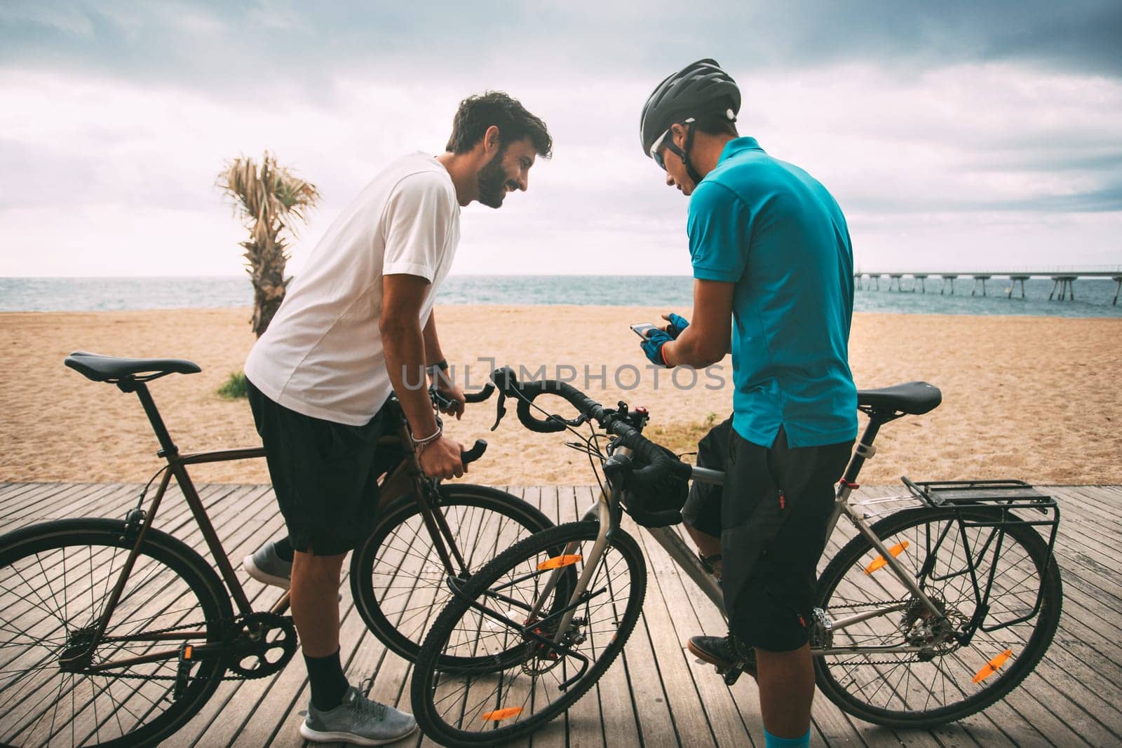 Cyclists on a bike on a wooden walkway on the beach with the sea and a bridge in the background looking at the mobile phone. Clear sky background with copyspace. sport concept. urban cycling concept. technology concept by Hoverstock