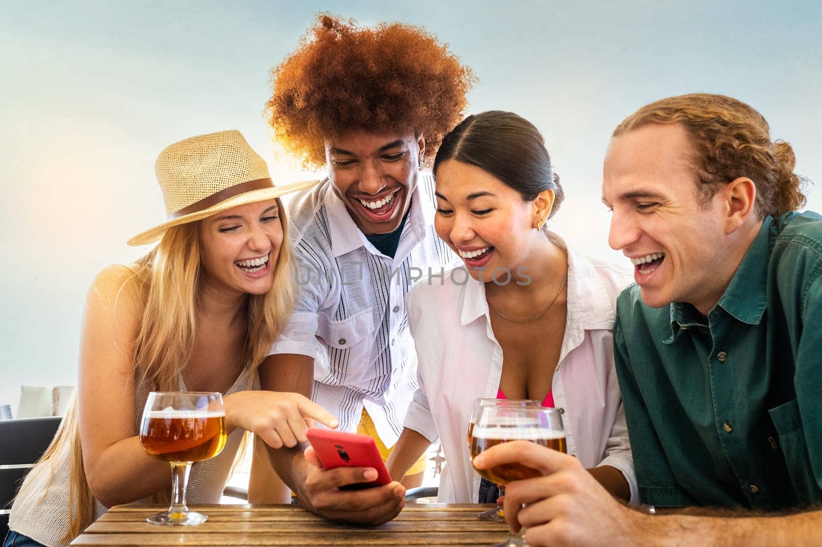Young African American man and his happy multiracial friends looking at mobile phone in a beach bar while drinking beer. Technology and leisure activity.