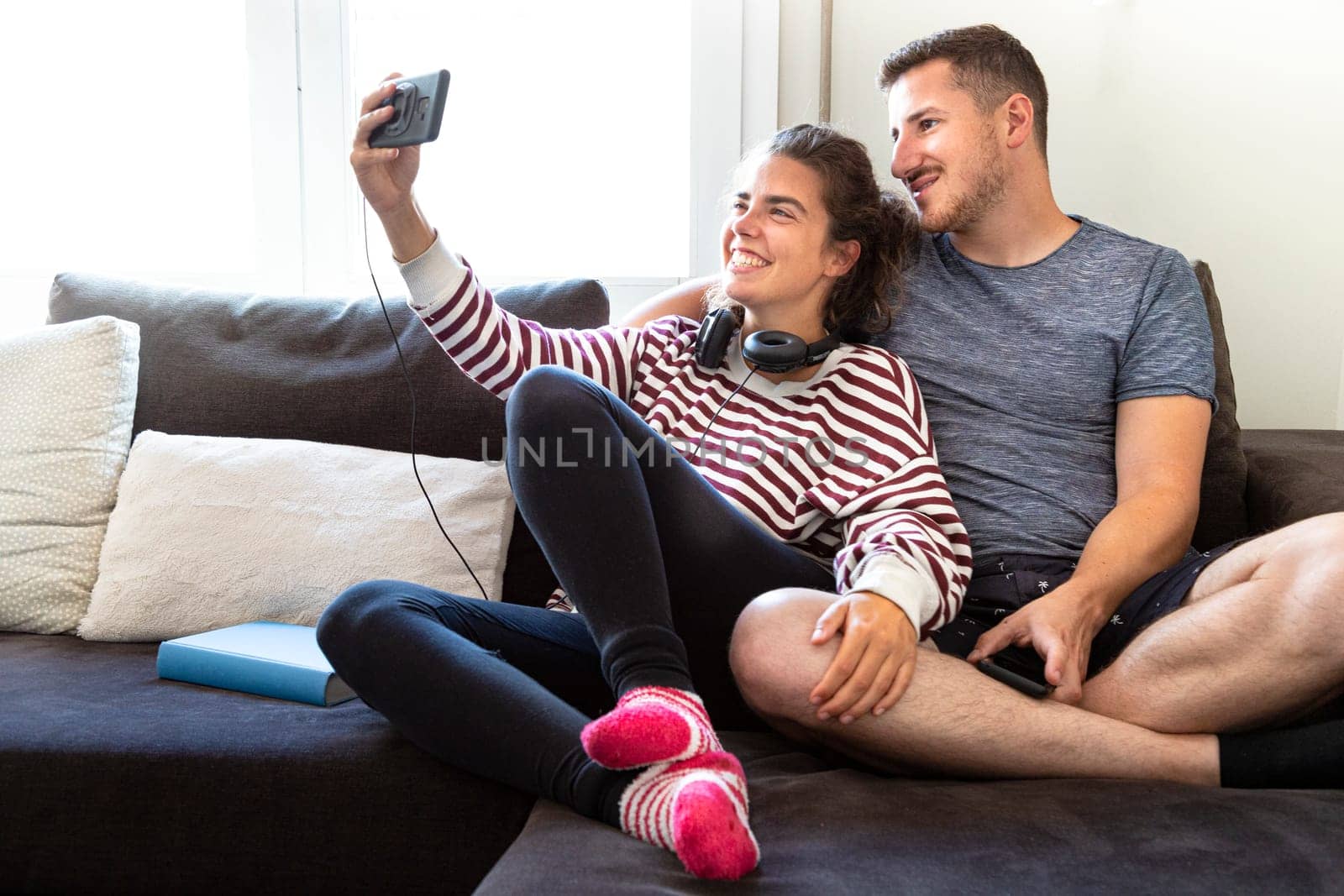 young couple sitting comfortably on a sofa making a smiling and happy selfie in front of a window in a very bright room by Hoverstock