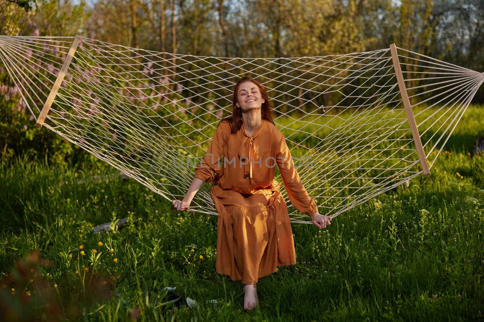 horizontal photo of a beautiful, red-haired woman lying in a hammock enjoying a rest in a long orange dress, on a warm summer day, smiling happily looking at the camera by Vichizh