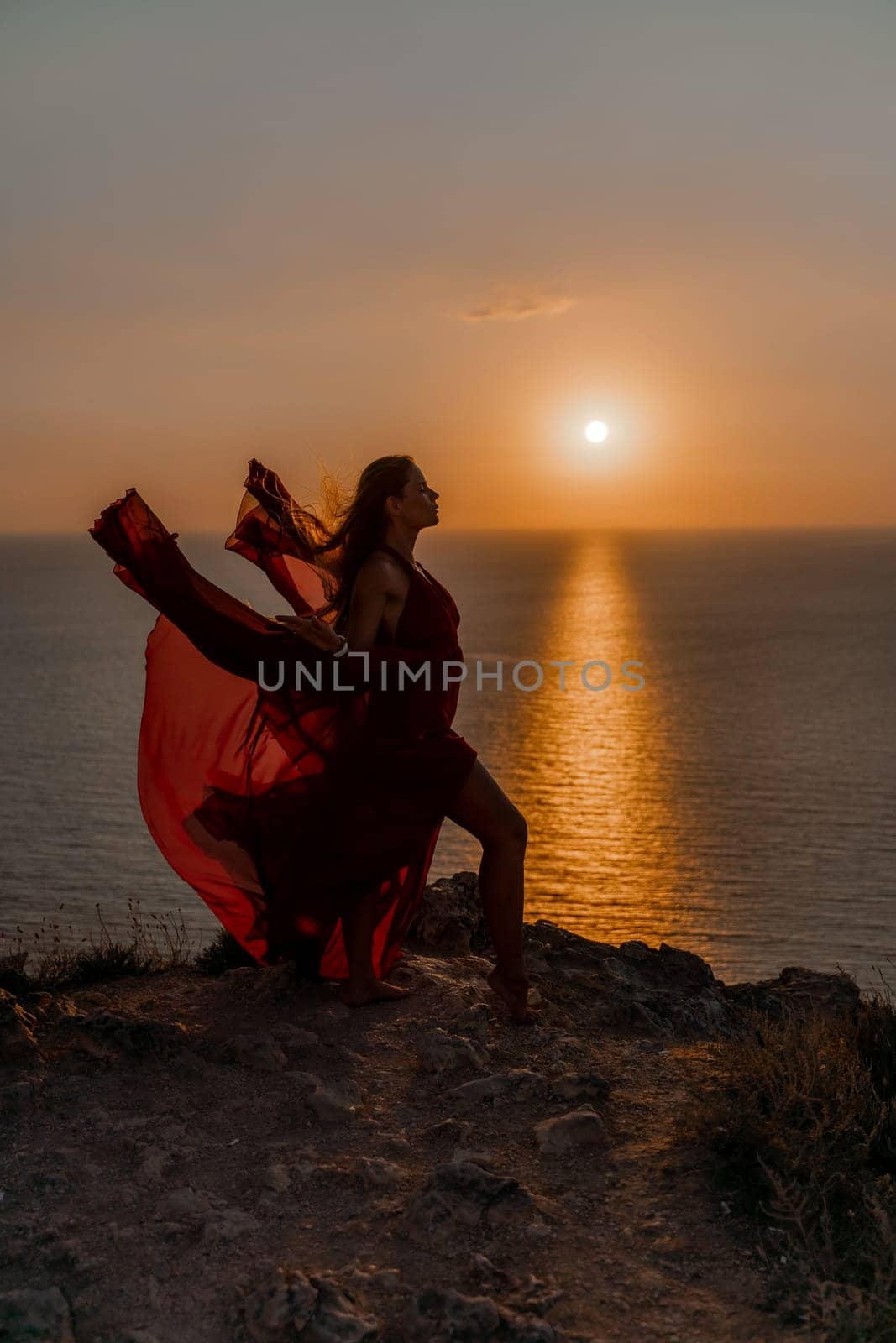 Woman sunset sea red dress, back view a happy beautiful sensual woman in a red long dress posing on a rock high above the sea on sunset. by Matiunina