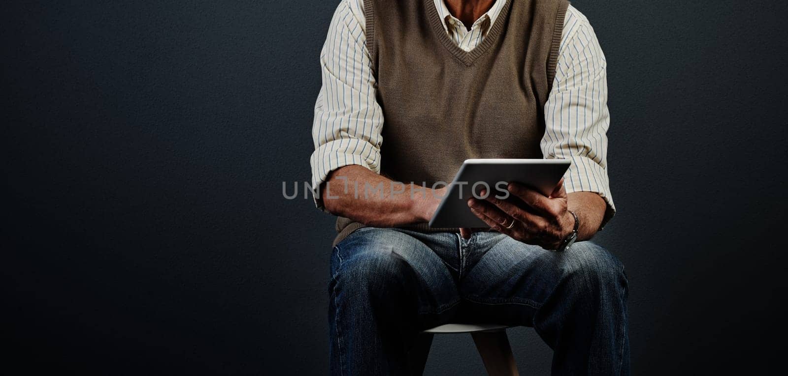 Updating his lifestyle. Studio shot of an unrecognizable man using a tablet while sitting on a wooden stool against a dark background