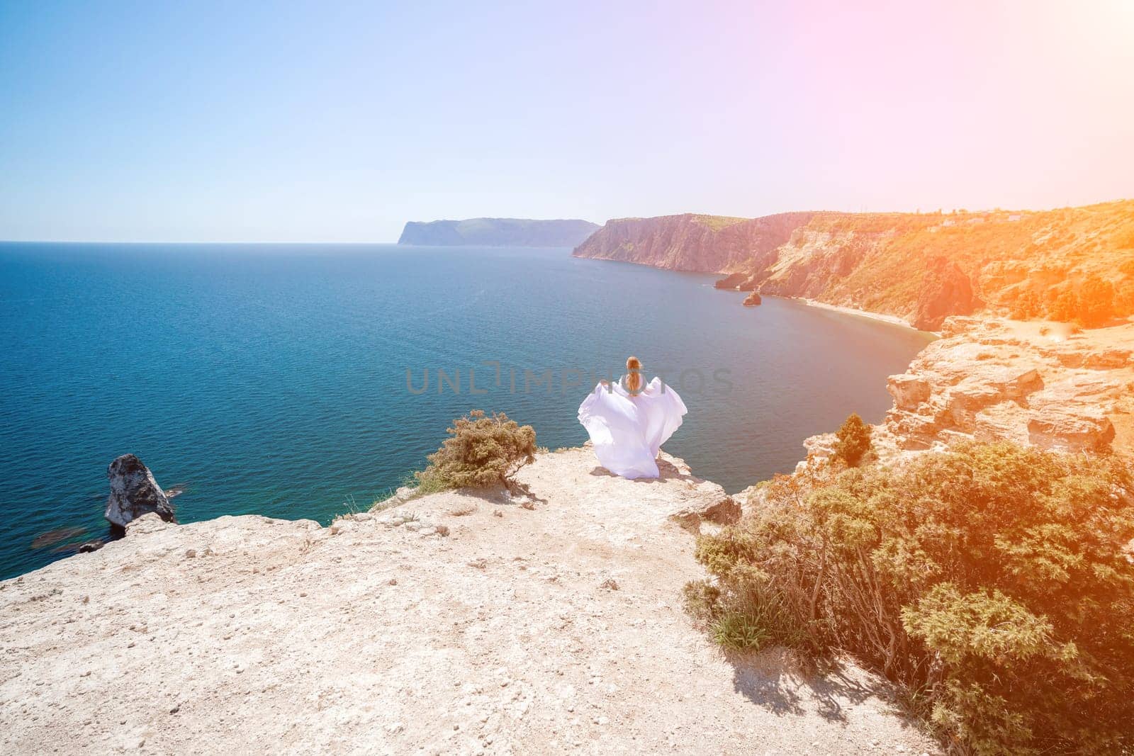 woman sea white dress. Blonde with long hair on a sunny seashore in a white flowing dress, rear view, silk fabric waving in the wind. Against the backdrop of the blue sky and mountains on the seashore