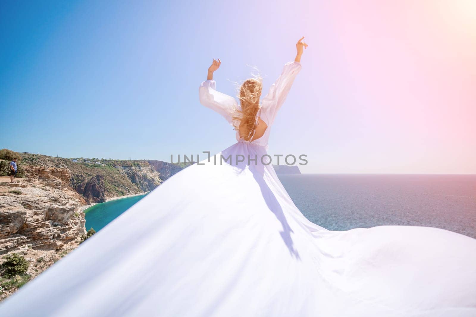 woman sea white dress. Blonde with long hair on a sunny seashore in a white flowing dress, rear view, silk fabric waving in the wind. Against the backdrop of the blue sky and mountains on the seashore. by Matiunina
