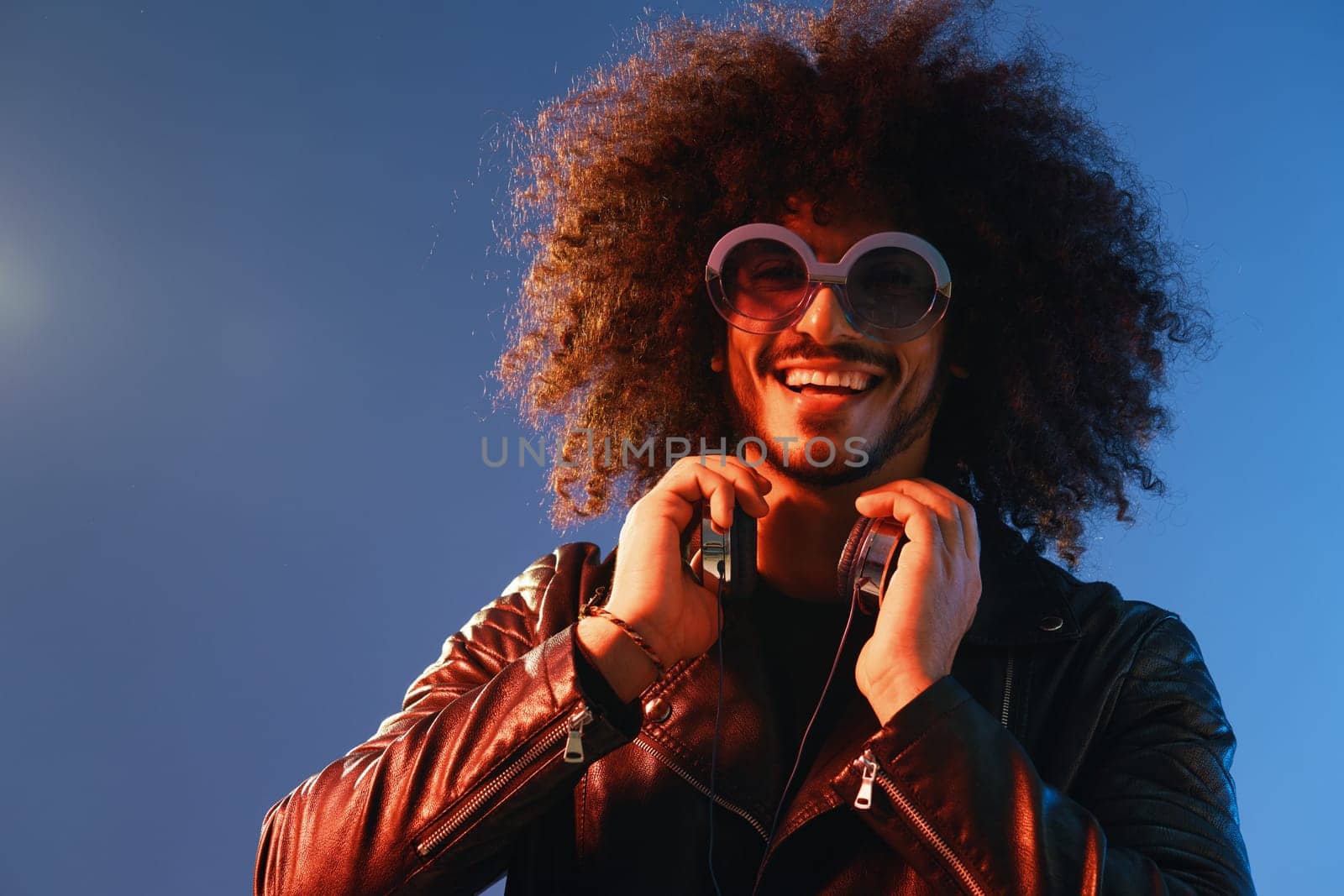 Portrait of a stylish man with curly hair with glasses and headphones on a blue background multinational, colored light, black leather jacket trend, modern concept. by SHOTPRIME