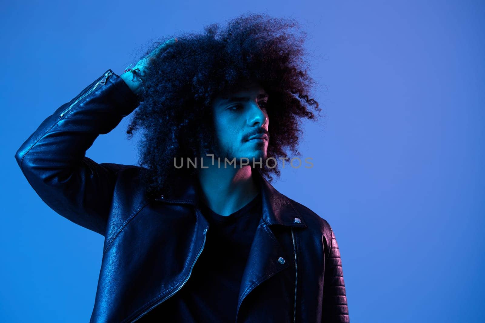 Portrait of fashion man with curly hair on blue background multinational, colored light, black leather jacket trend, modern concept. by SHOTPRIME