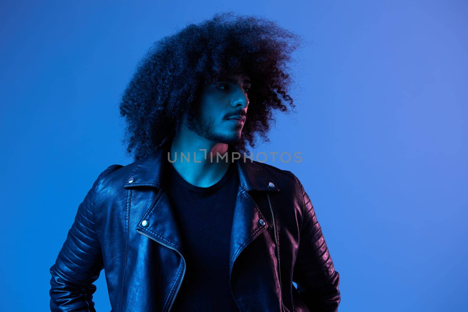 Portrait of fashion man with curly hair on blue background multinational, colored light, black leather jacket trend, modern concept. by SHOTPRIME