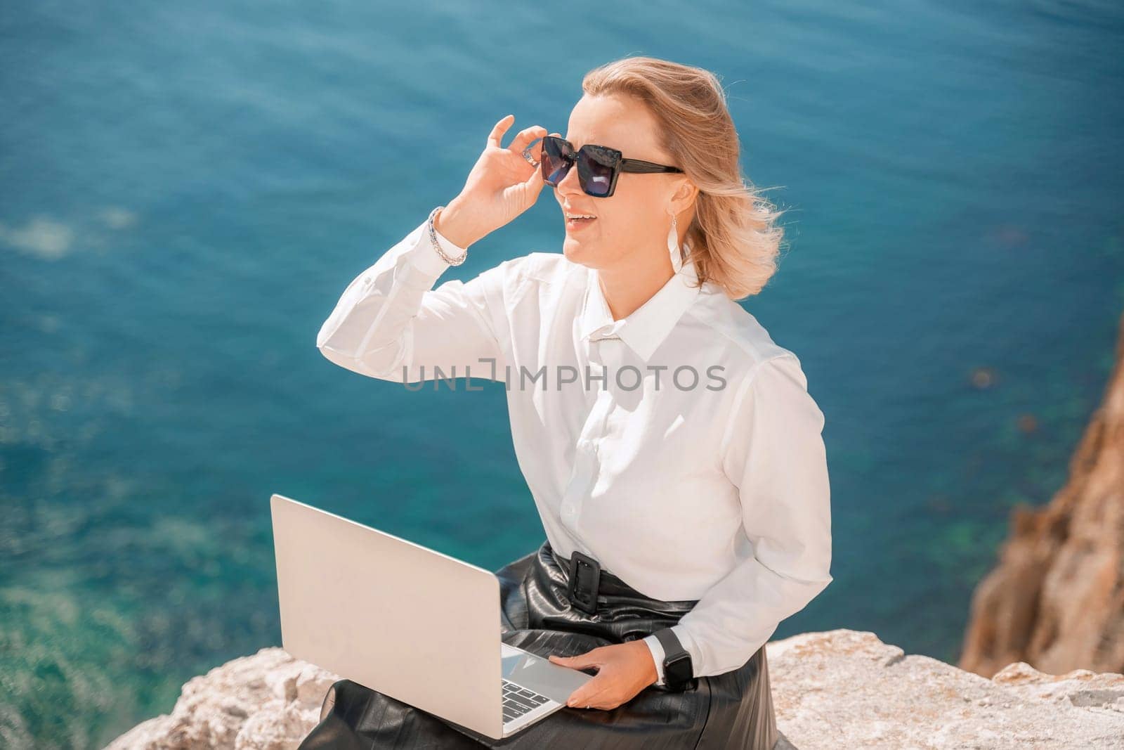 Business woman on nature in white shirt and black skirt. She works with an iPad in the open air with a beautiful view of the sea. The concept of remote work. by Matiunina