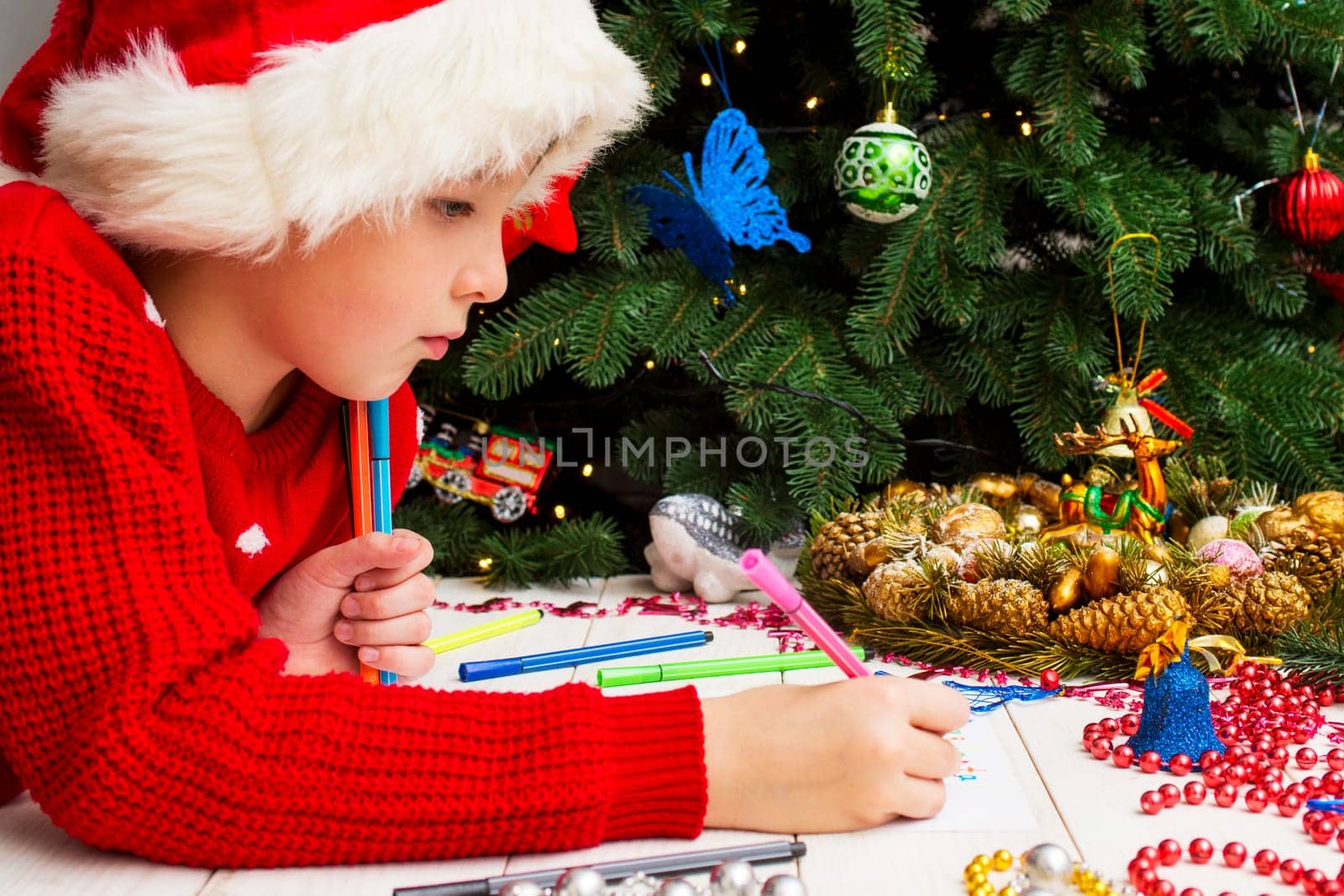 Little child writes letter to Santa. Cute boy in Santa hat writes a letter to Santa near the Christmas tree.