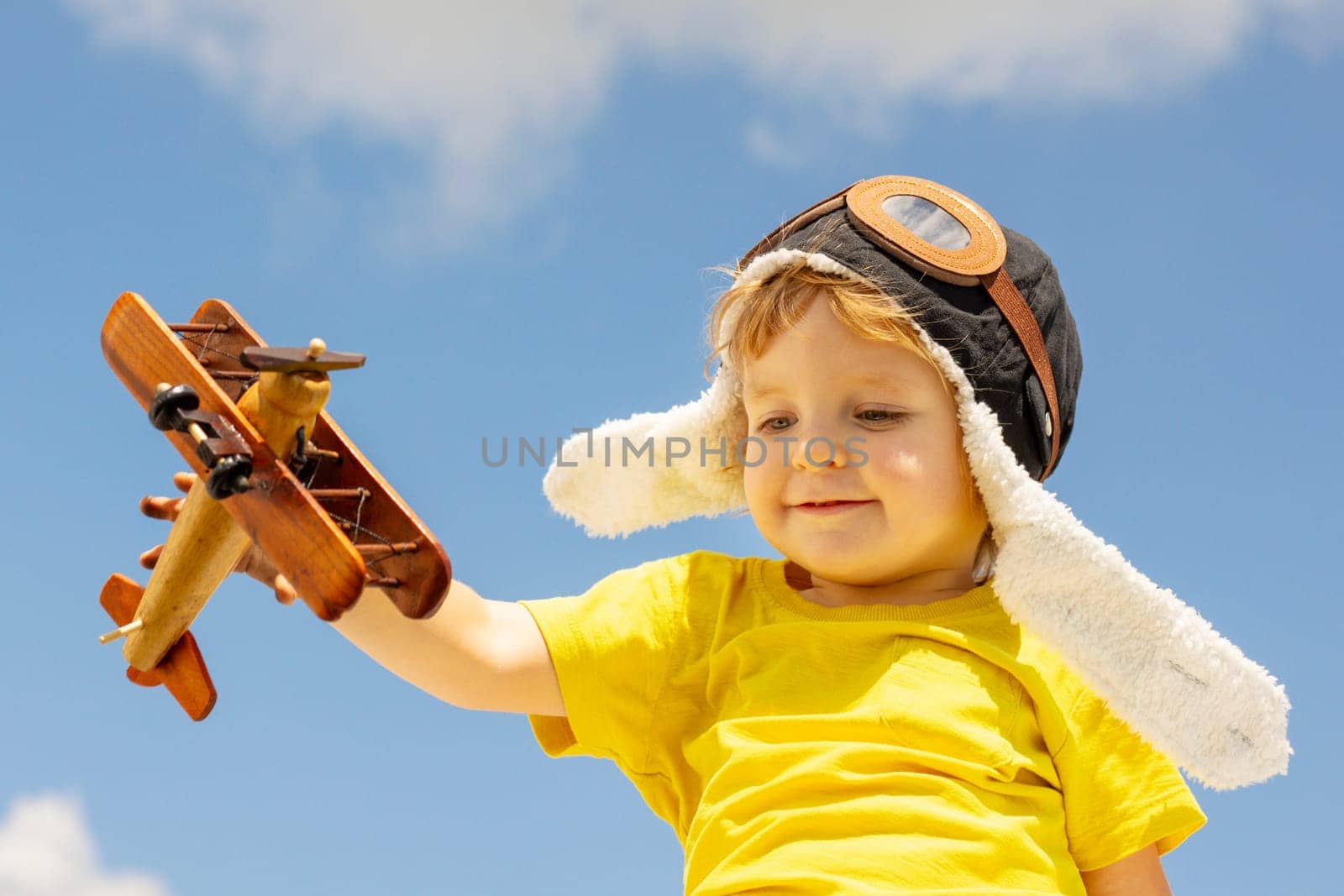 Happy child boy playing with wooden toy airplane on blue sky background with clouds.