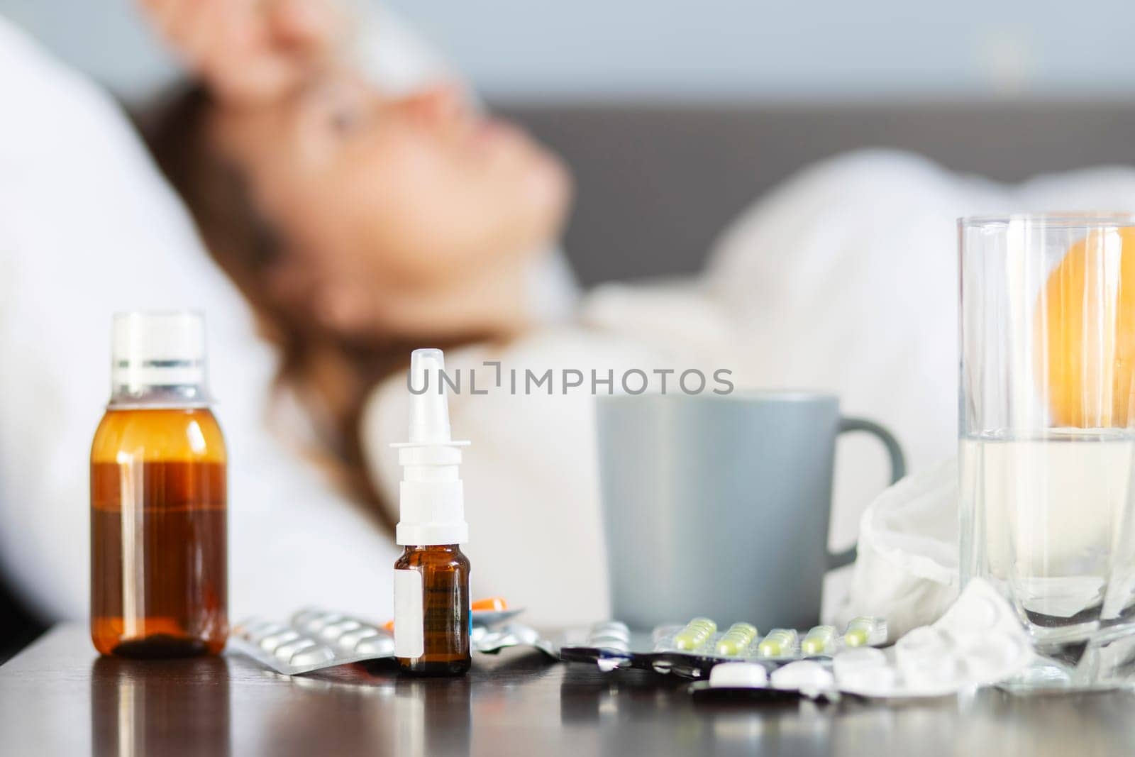 Sick young woman covered with a blanket lying in bed with high fever and a flu. A lot of medicines on table in bedroom.