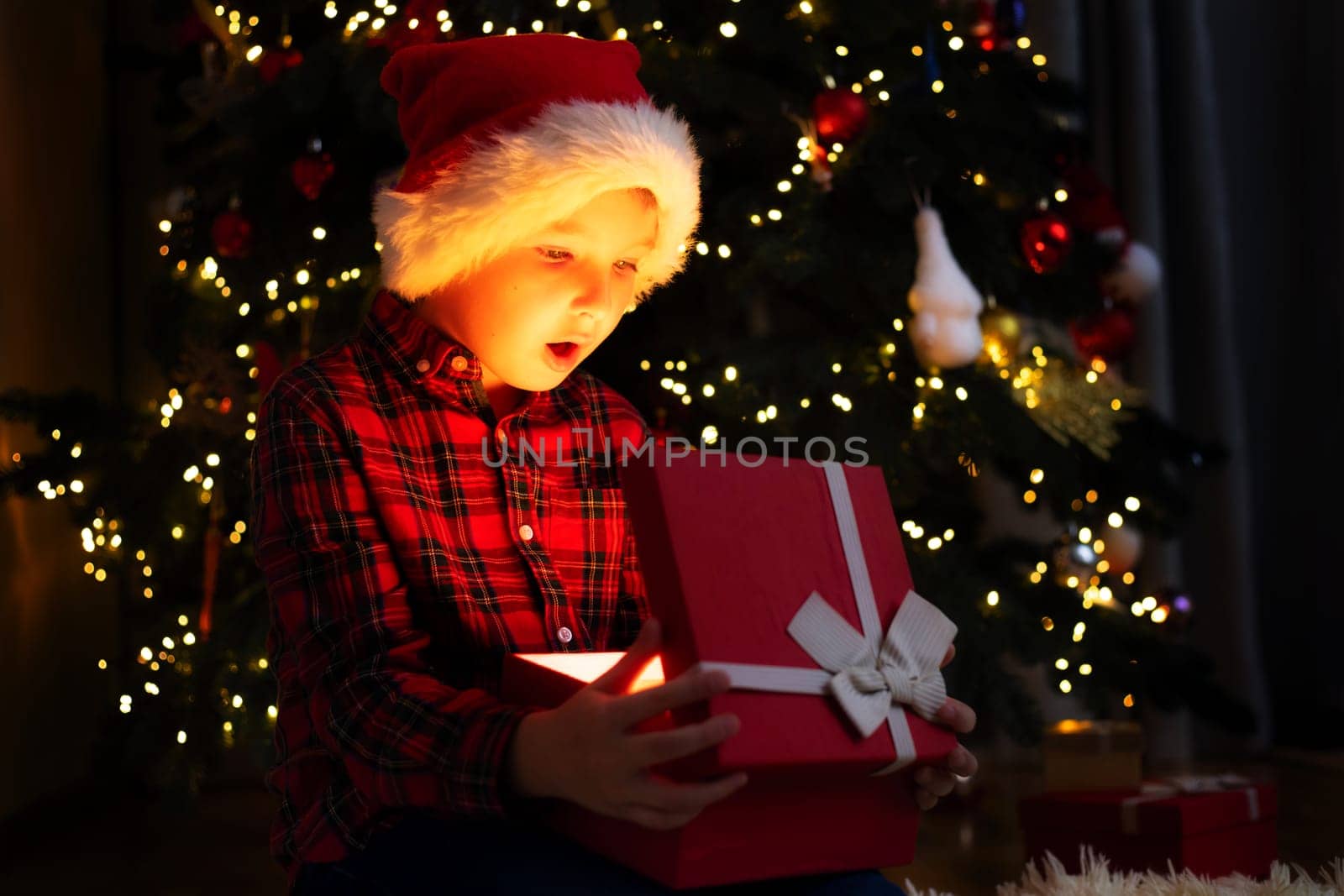 Cute little boy opening magic Christmas gift by a Christmas tree in cozy living room in winter. Merry Christmas