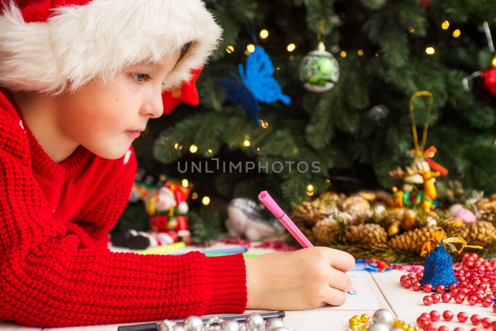 Little child is writing letter to Santa Claus at table. A kid in Santa hat is writing wishlist near the Christmas tree.
