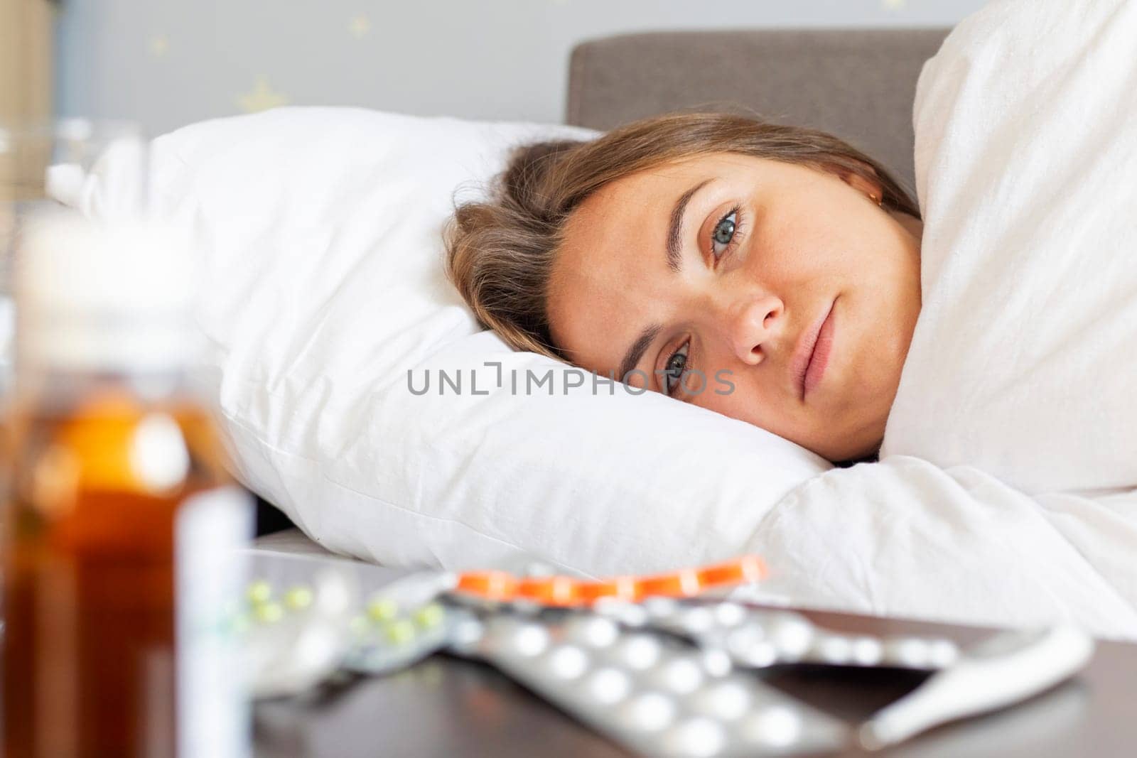 Sick young woman covered with a blanket lying in bed with high fever and a flu. A lot of medicines on table in bedroom.