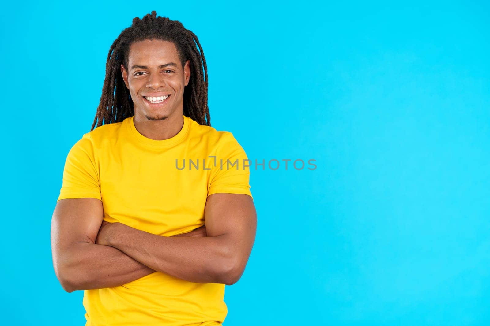 Happy latin man with dreadlocks standing with arms crossed in studio with blue background