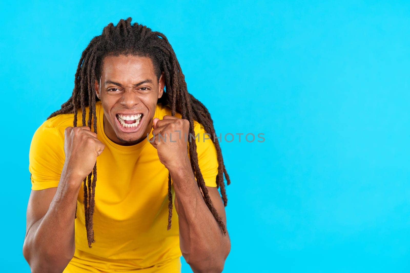 Happy hispanic man with dreadlocks gesturing with fists while celebrating in studio with blue background