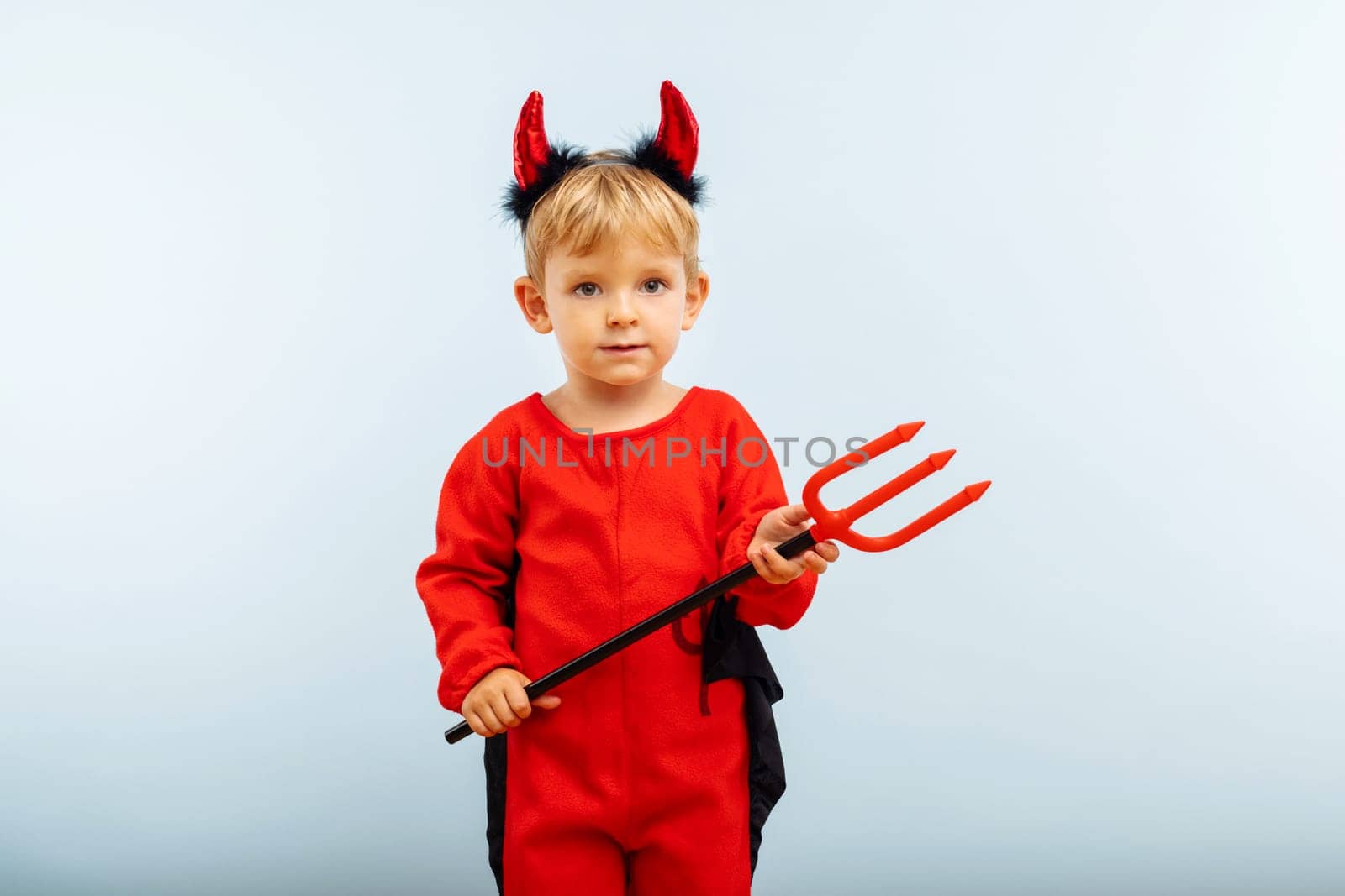 Happy Halloween. Cute little boy in devil halloween costume with horns and trident on light blue background.