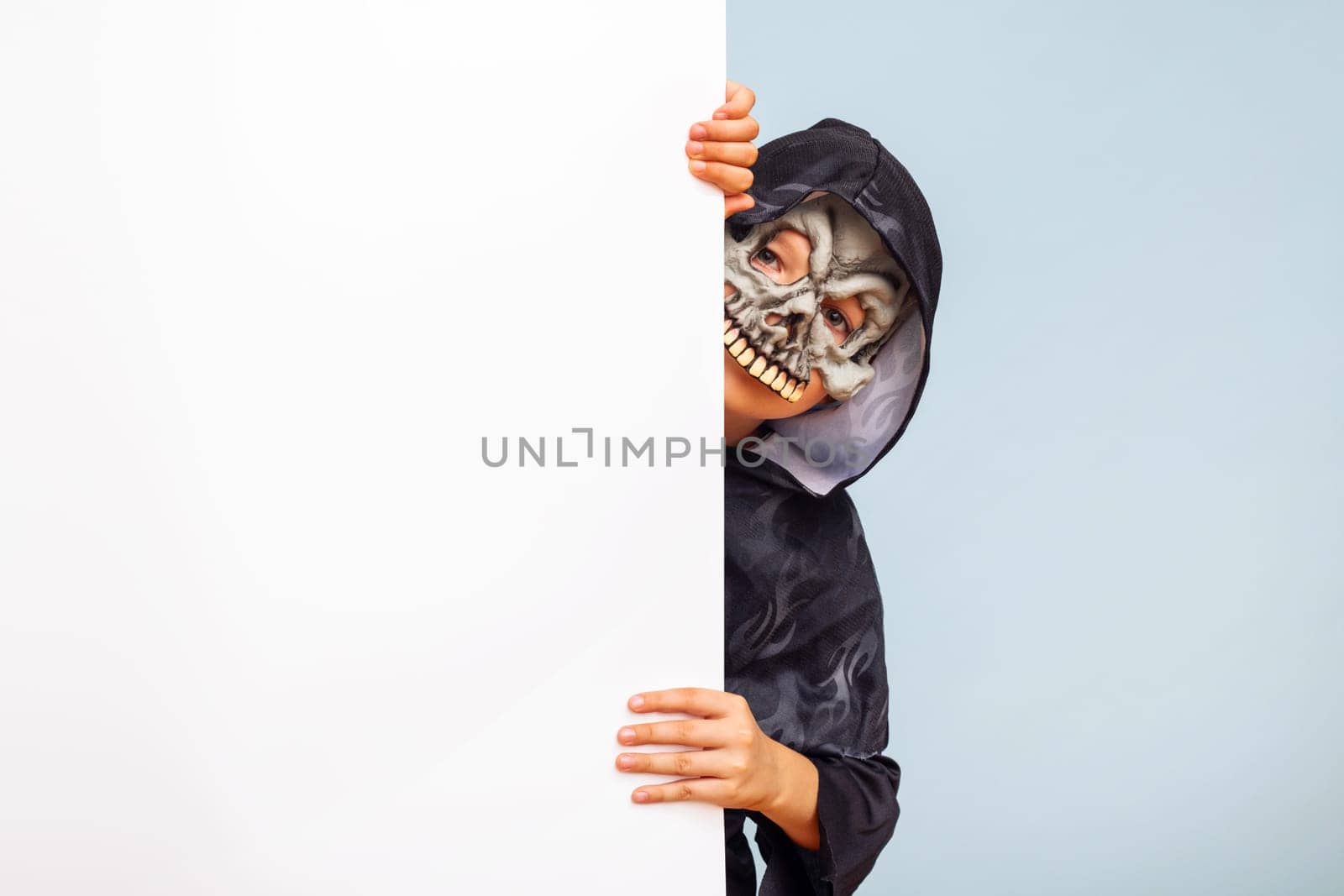 Happy Halloween. Cute little boy in a costume hiding behind white banner blank. Happy kid holding cardboard background with copy space.