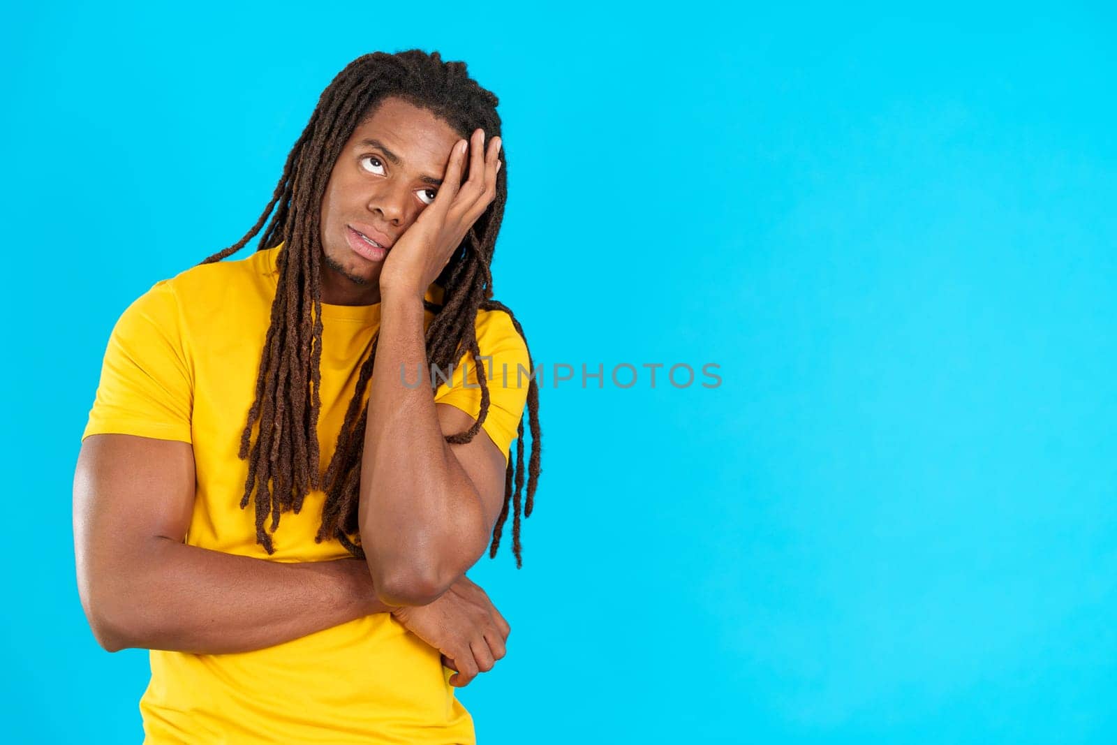 Bored latin man with dreadlocks standing and looking up in studio with blue background