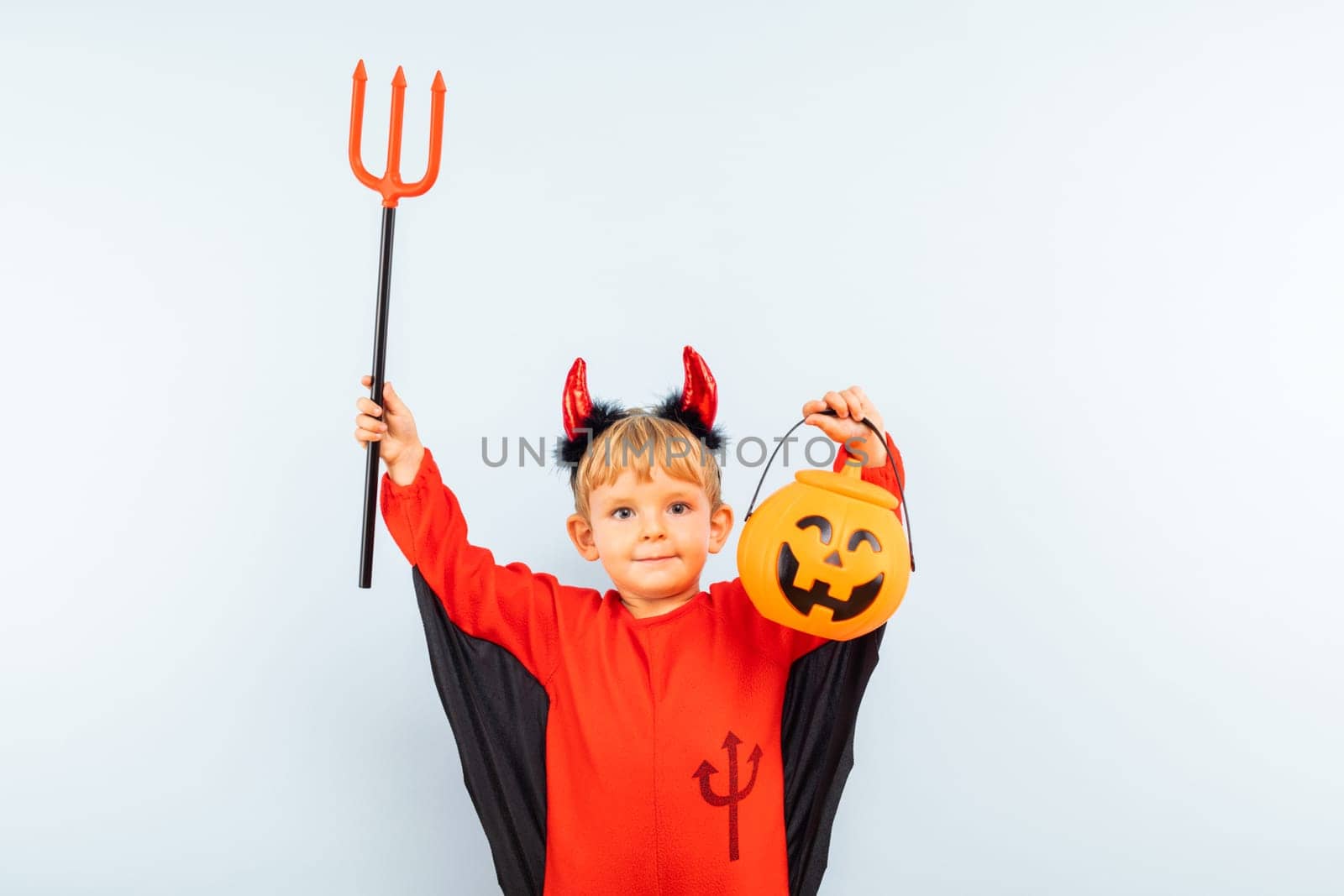 Cute boy in devil halloween costume with trident and pumpkin basket by andreyz