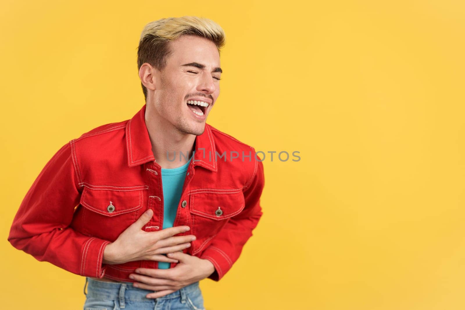 Stylish gay man in colorful clothes laughing while standing in studio with yellow background