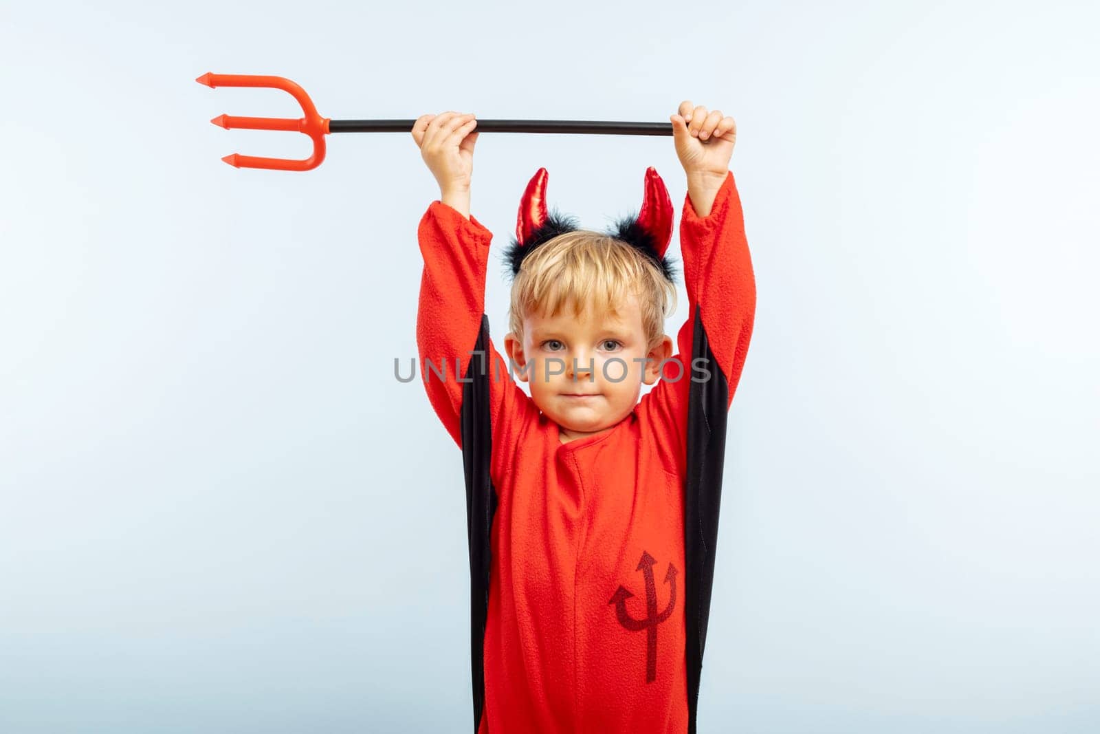 Happy Halloween. Cute little boy in devil halloween costume with horns and trident on light blue background.